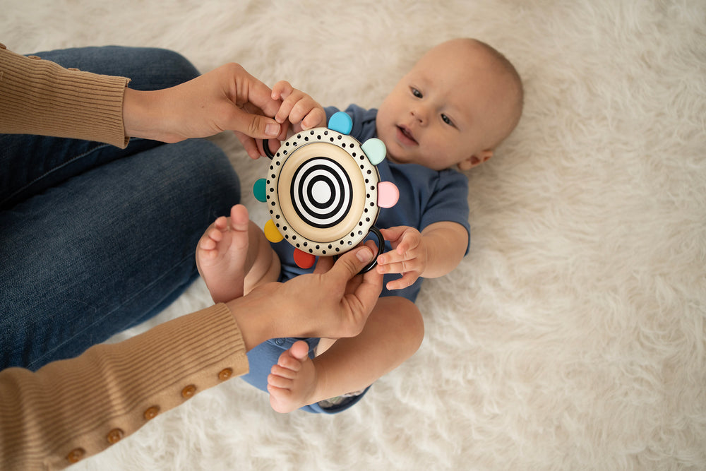 Baby lying on a soft rug, grasping a high-contrast circular mirror toy with colorful tabs, supported by an adult's hands.