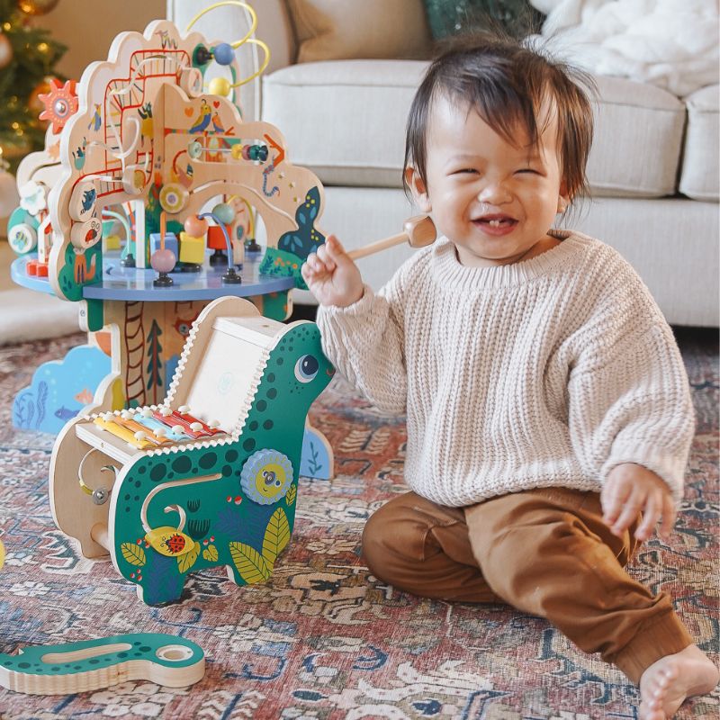 Little boy smiling while holding a wooden mallet seated next to a dinosaur wood musical toy and a wooden activity center in a living room scene. 