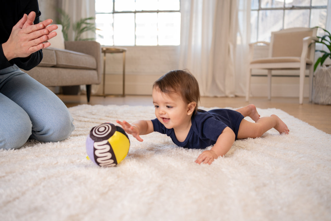 Baby laying on their tummy reaching for a ball toy while mom claps