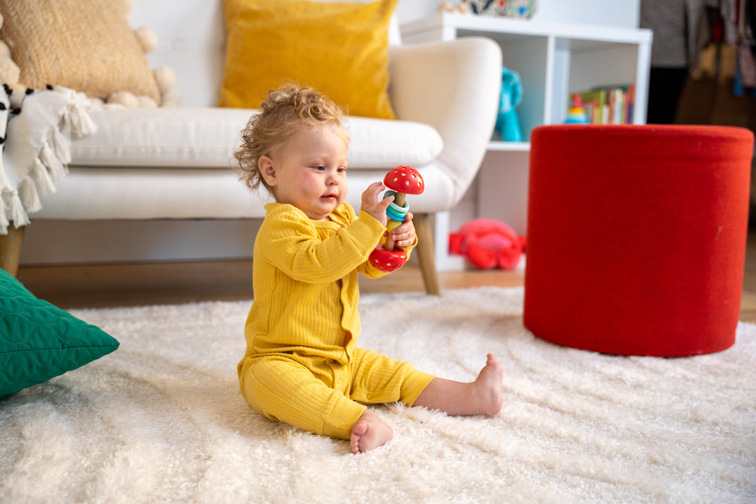 Baby girl sitting on living room floor playing with a barbell rattle.