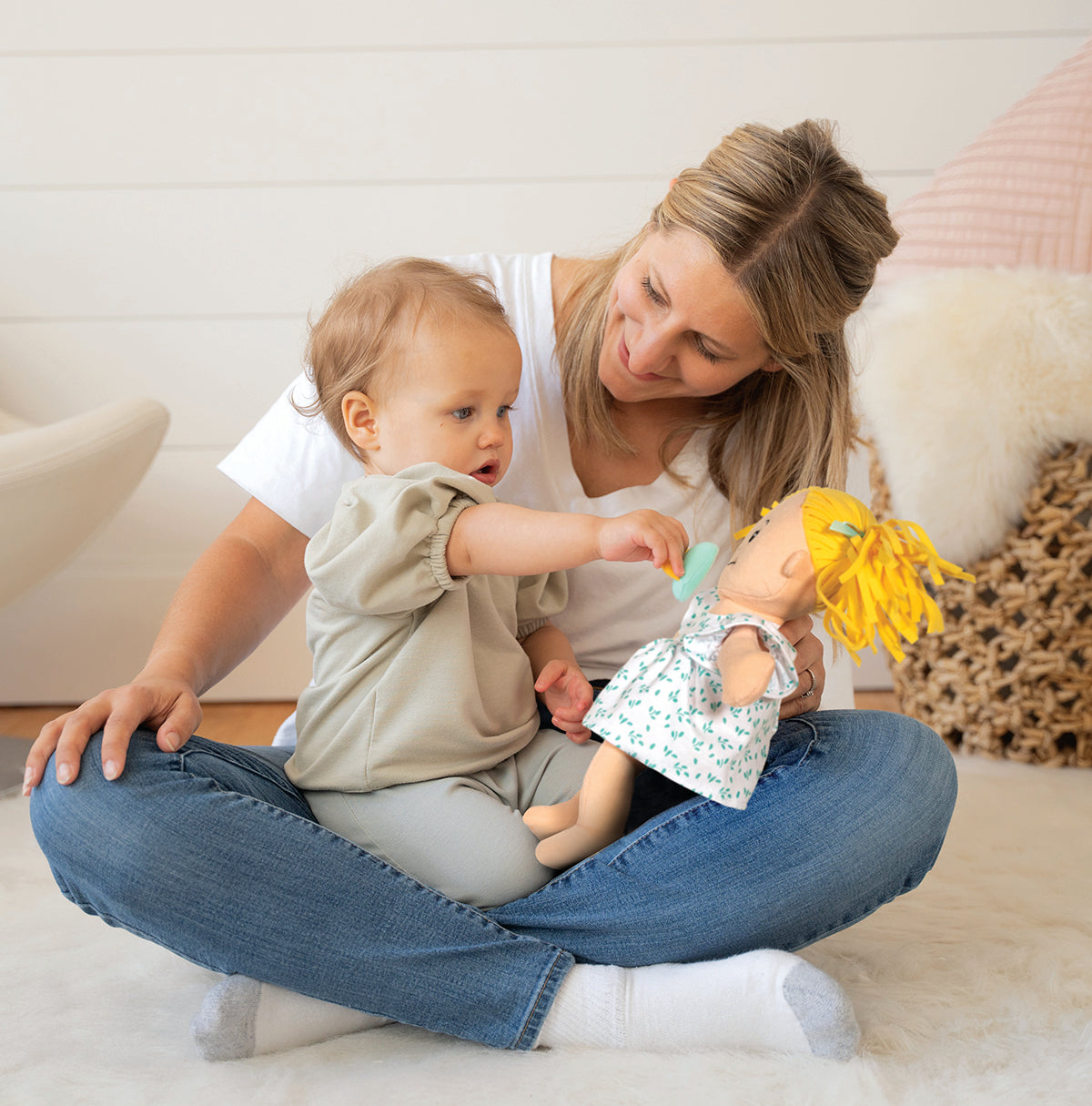 Mom and toddler age daughter sitting in a room scene. Daughter is sitting on mom's lap playing with a soft baby doll, placing a pacifier on the dolls face, while mom smiles at her daughter.