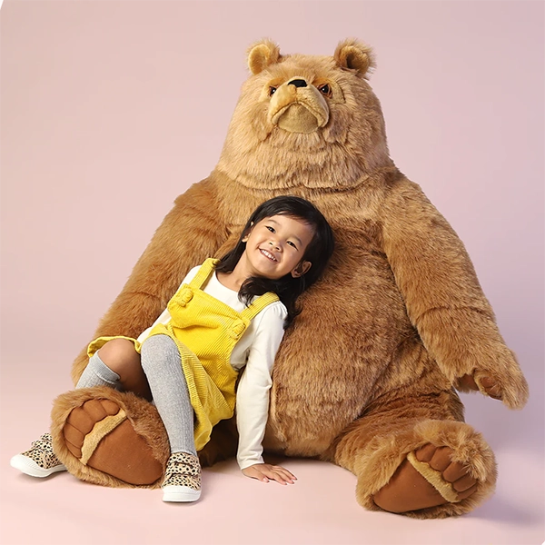 Smiling young girl sitting against a giant plush bear on a pink background, wearing a yellow dress and gray tights.